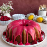 The lemon raspberry bundt cake with a raspberry glaze on a plate. There are fresh raspberries and dessert plates in the background