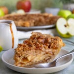 A slice of apple caramel crumble pie on a dessert plate with a spoon. The rest of the pie is behind the slice.
