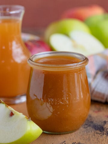 An 8-ounce jar filled with apple cider caramel sauce. The jar is surrounded by fresh apples and there is a small jar of apple cider behind jar of caramel sauce.