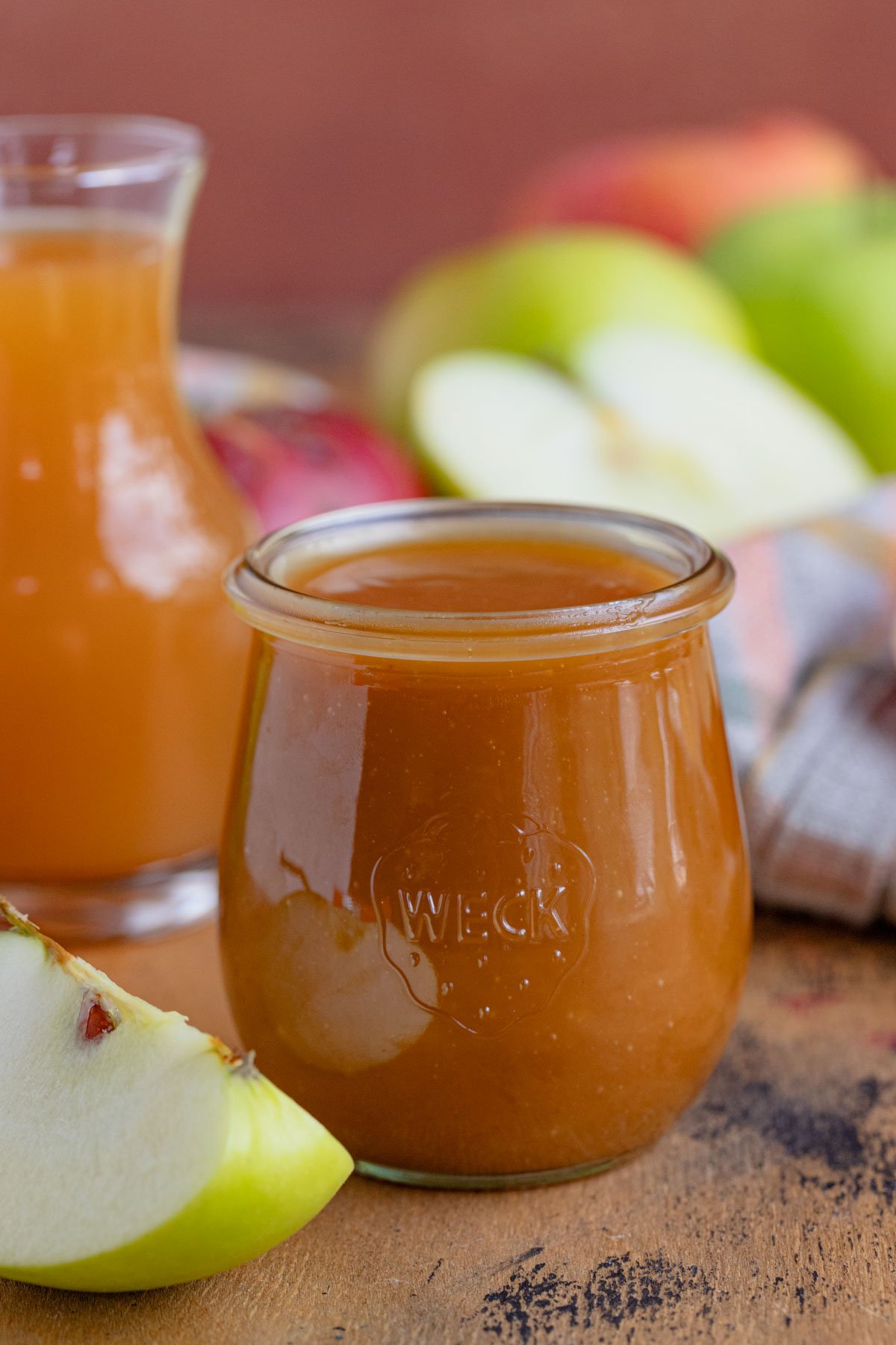 An 8-ounce jar filled with apple cider caramel sauce.  The jar is surrounded by fresh apples and there is a small jar of apple cider behind jar of caramel sauce.