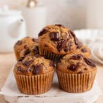 Four chocolate chip muffins are arranged on a white wooden board. The muffins have a golden-brown color with visible chunks of melted chocolate. In the background, there is a blurred white teapot, cups, and a glass bottle.