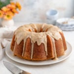 Sour cream pound cake with icing drizzled over the top sits on a white plate. A knife with a wooden handle lies nearby on a white surface. In the background, there are stacked plates and a small vase with orange flowers.