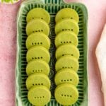 A rectangular green tray with 2 rows of green matcha shortbread cookies. There are 2 cups of tea and a bouquet of flowers nearby.