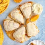 Six triangle-shaped orange scones topped with an orange glaze arranged on a round board, surrounded by fresh oranges,.