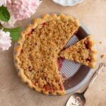 A round pie with crumb topping with one slice removed. A pie server with crumbs rests beside the dish, and pink flowers and an empty white plate are in the background.