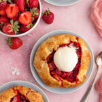 Three mini strawberry rhubarb galettes are on light blue plates, with one topped with a scoop of vanilla ice cream. A bowl of fresh strawberries and a spoon are also visible.