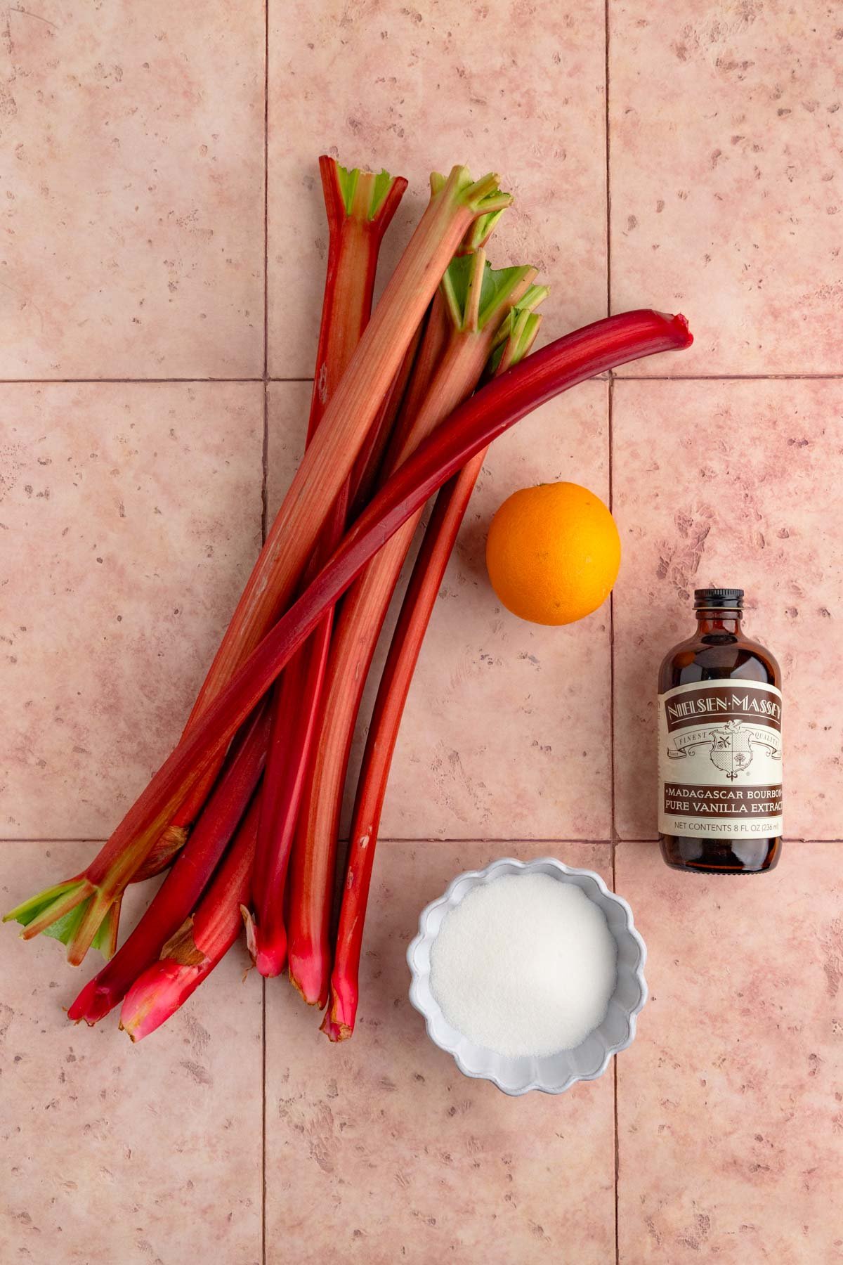 Rhubarb compote ingredients - fresh rhubarb, an orange, sugar, and pure vanilla extract laid out on a pink background