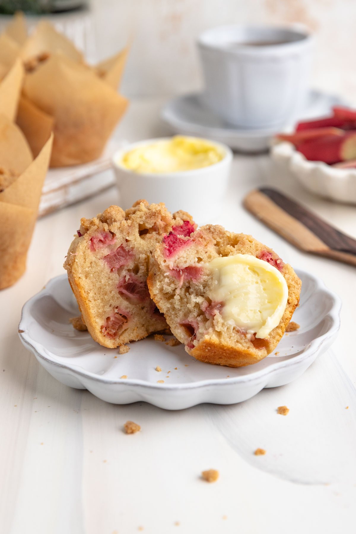A rhubarb muffin cut in half on a small plate. One half of the muffin is schmeared with butter. There is a small container of butter behind and more muffins to the left.