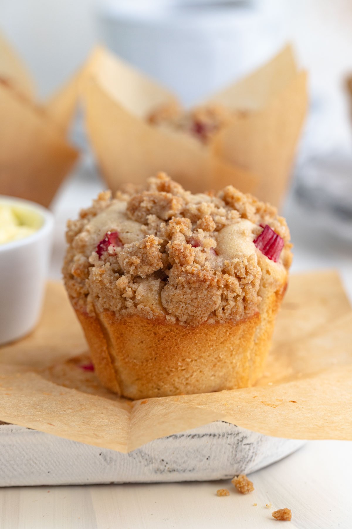 rhubarb muffin on a piece of brown parchment paper with a small dish of butter to the left and more muffins in the background