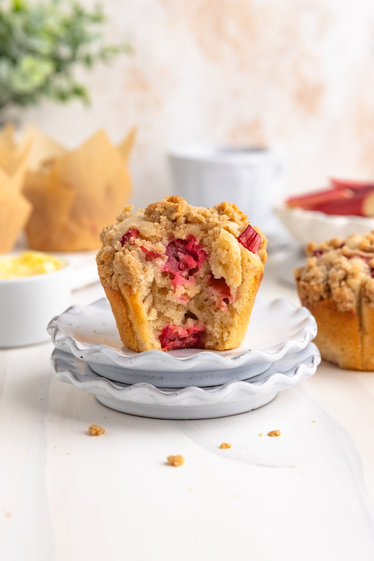 Two small plates stacked with a single muffin on top. There is a muffin behind on the right, and there are more muffins in the back. There is a small dish of butter to the left.