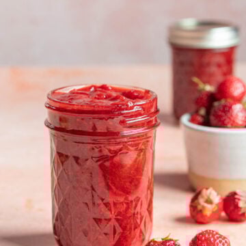 A glass jar filled with strawberry jam is placed on a light pink surface. Fresh strawberries are scattered nearby, with another jar and a small bowl of strawberries in the background.