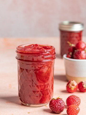 A glass jar filled with strawberry jam is placed on a light pink surface. Fresh strawberries are scattered nearby, with another jar and a small bowl of strawberries in the background.