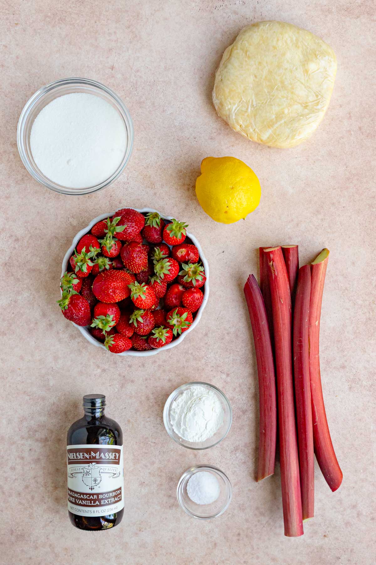 Strawberry rhubarb pie ingredients on a board - granulated sugar, a lemon, pie dough wrapped in plastic, fresh rhubarb, cornstarch, kosher salt, pure vanilla extract, and fresh strawberries