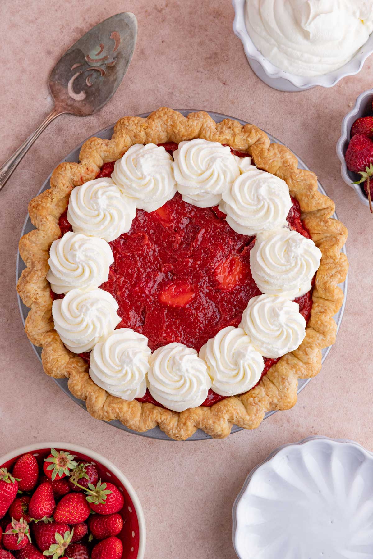 Strawberry Rhubarb Pie topped with swirls of whipped cream.  There are two bowl of strawberries on either side of the pie and a white dessert plate on the bottom right