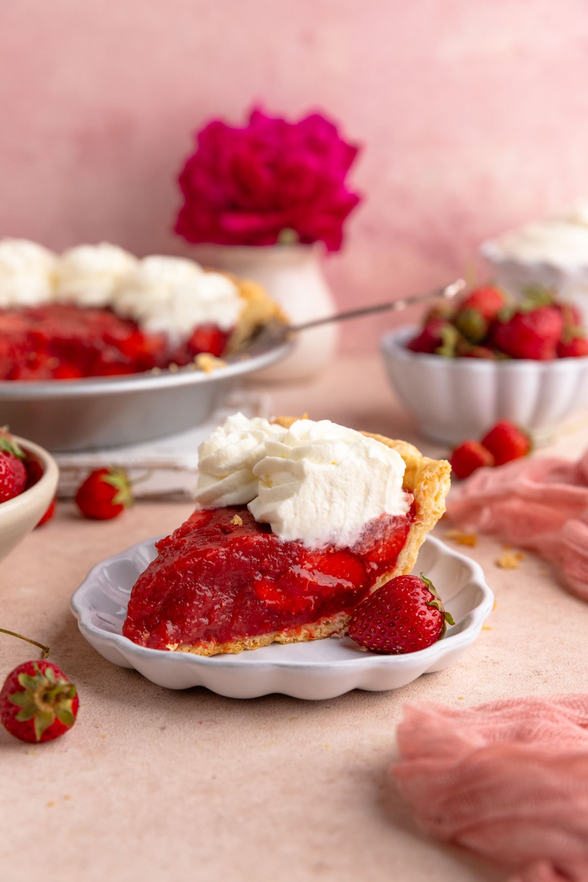 A slice of rhubarb pie topped with whipped cream on a dessert plate.  The rest of the pie is in the background along with a small bowl of fresh strawberries