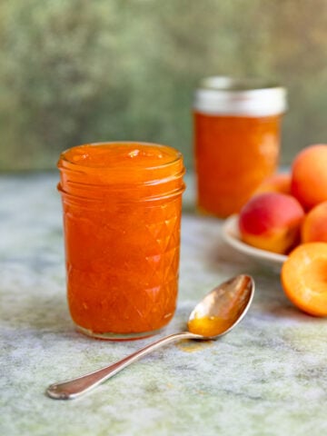Apricot jam in a pint jar with a half apricot on the left side of the jar. There is a spoon with apricot jam in front of the jar. The jam is bright orange.