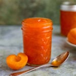 Apricot jam in a pint jar with a half apricot on the left side of the jar. There is a spoon with apricot jam in front of the jar. The jam is bright orange.