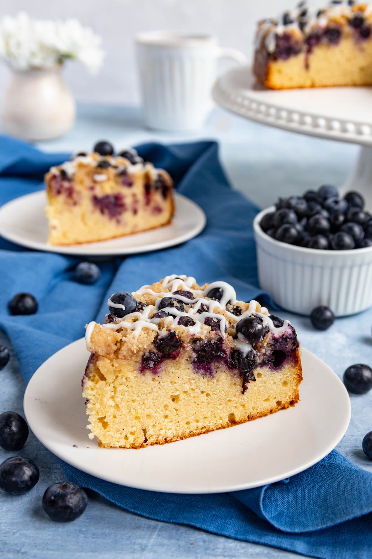 A slice  of blueberry coffee cake on a small plate with a second slice on a plate in the backgrand.  The rest of the cake is on a cake stand