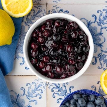 A white bowl filled with blueberry compote. Nearby are a bowl of fresh blueberries, a blue napkin, and halved lemons.