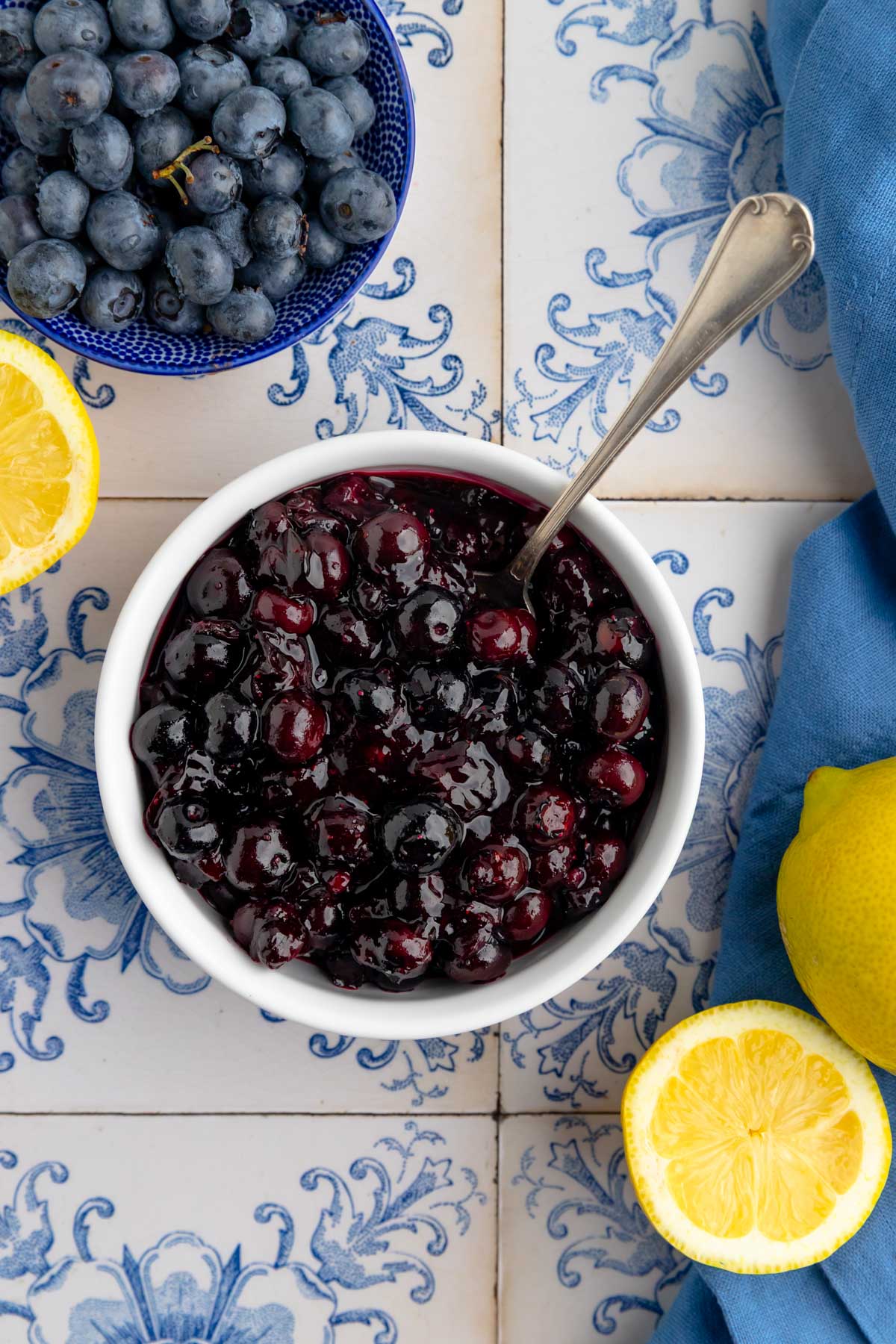 A white bowl filled with blueberry compote with a spoon in the bowl. Nearby are a bowl of fresh blueberries, a blue napkin, and halved lemons.