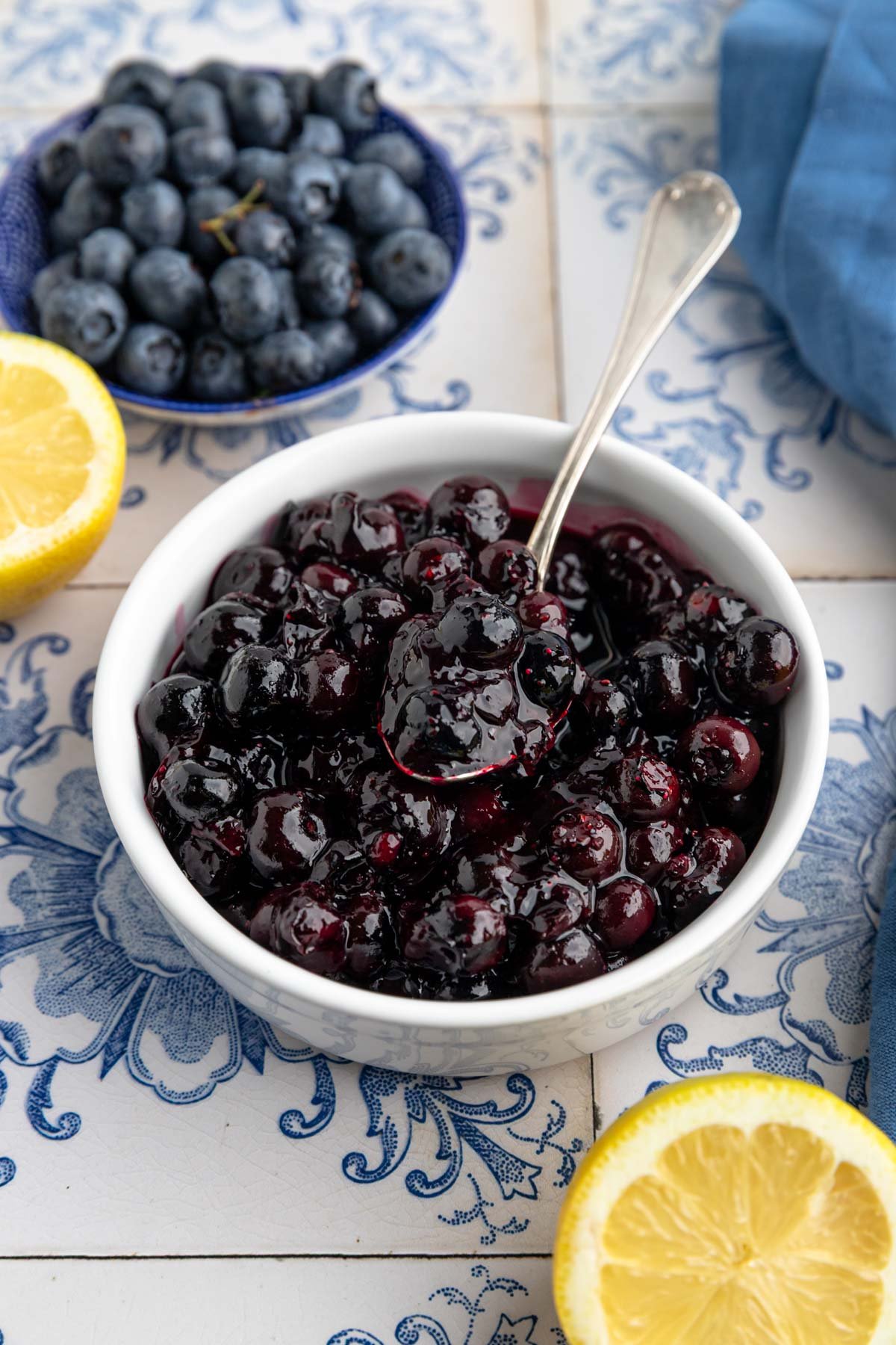 A white bowl filled with blueberry compote with a spoon in the bowl. Nearby are a bowl of fresh blueberries, a blue napkin, and halved lemons.