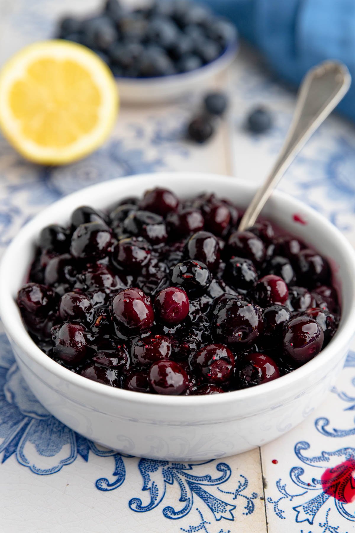 A white bowl filled with blueberry compote with a spoon in the bowl. Nearby are a bowl of fresh blueberries, a blue napkin, and halved lemons.