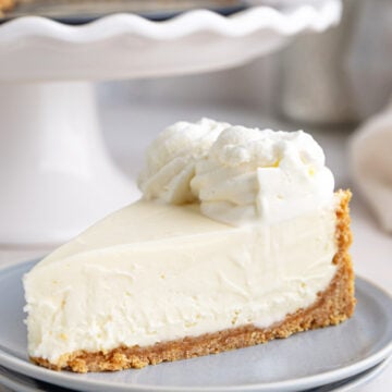 A slice of no-bake cheesecake with a graham cracker crust and whipped cream on top sits on a light gray plate, with the rest of the cheesecake on a cake stand in the background.