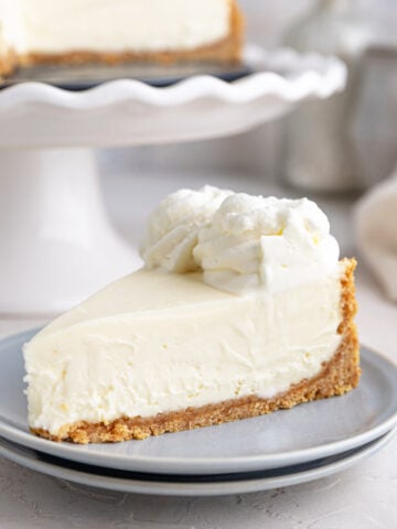 A slice of no-bake cheesecake with a graham cracker crust and whipped cream on top sits on a light gray plate, with the rest of the cheesecake on a cake stand in the background.