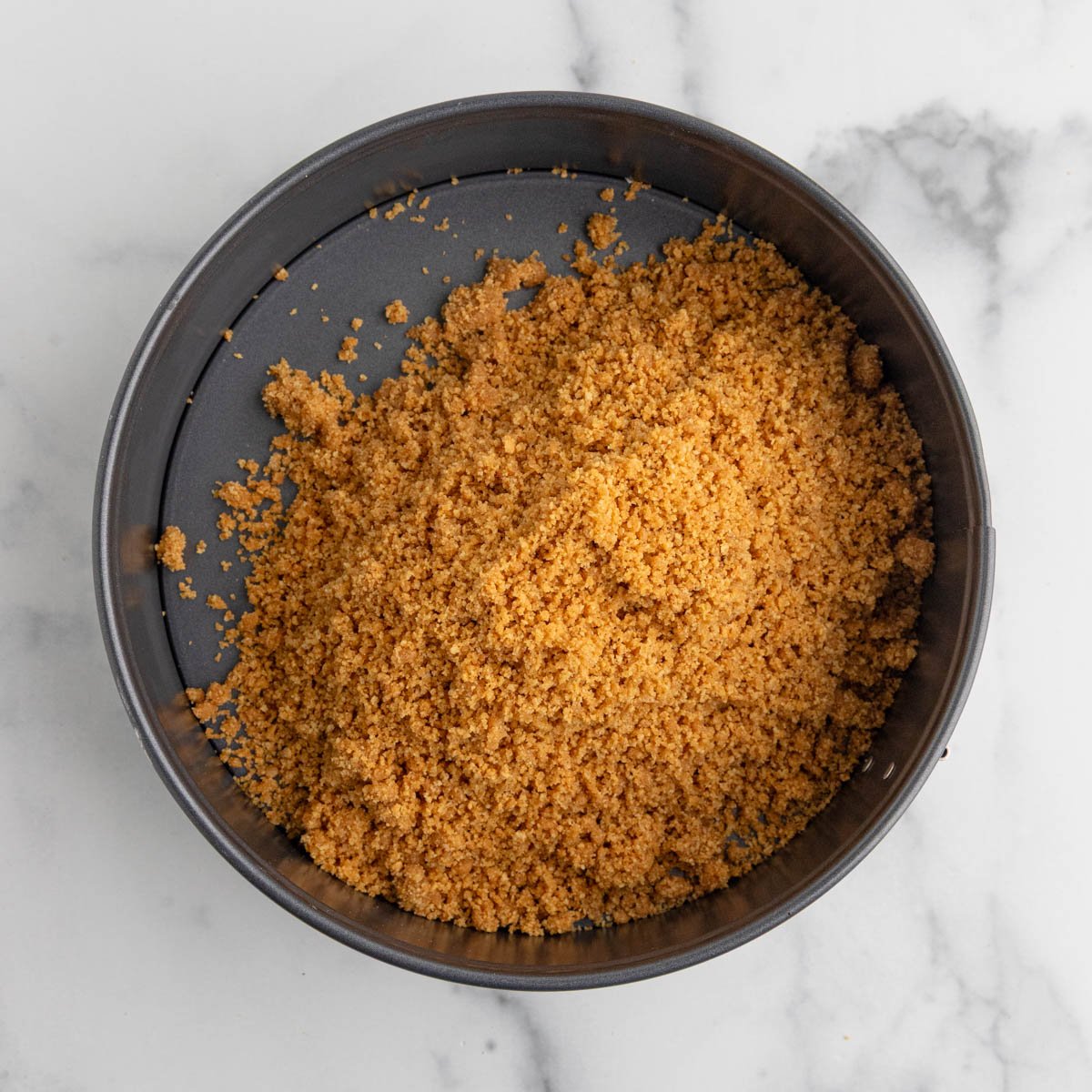 A springform pan on a marble surface filled with graham cracker crumbs.