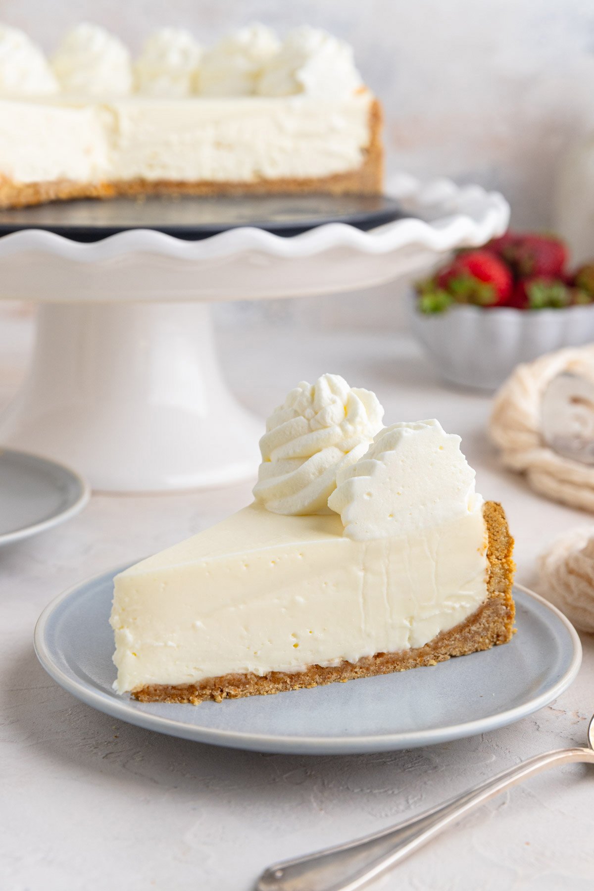 A slice of no-bake cheesecake with a graham cracker crust and whipped cream on top sits on a light gray plate, with the rest of the cheesecake on a cake stand in the background.