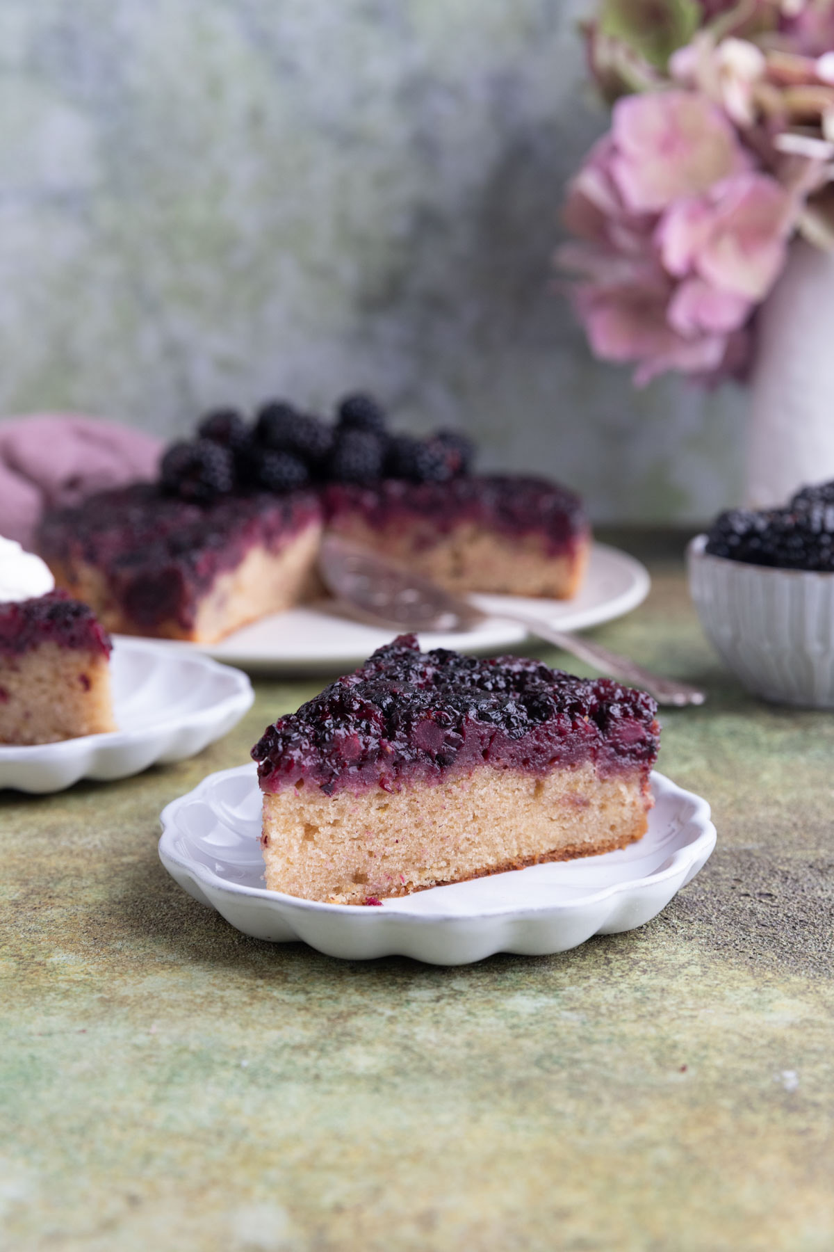 A slice of blackberry upside down cake on a small white plate with the rest of the cake and a bowl of fresh blackberries in the background
