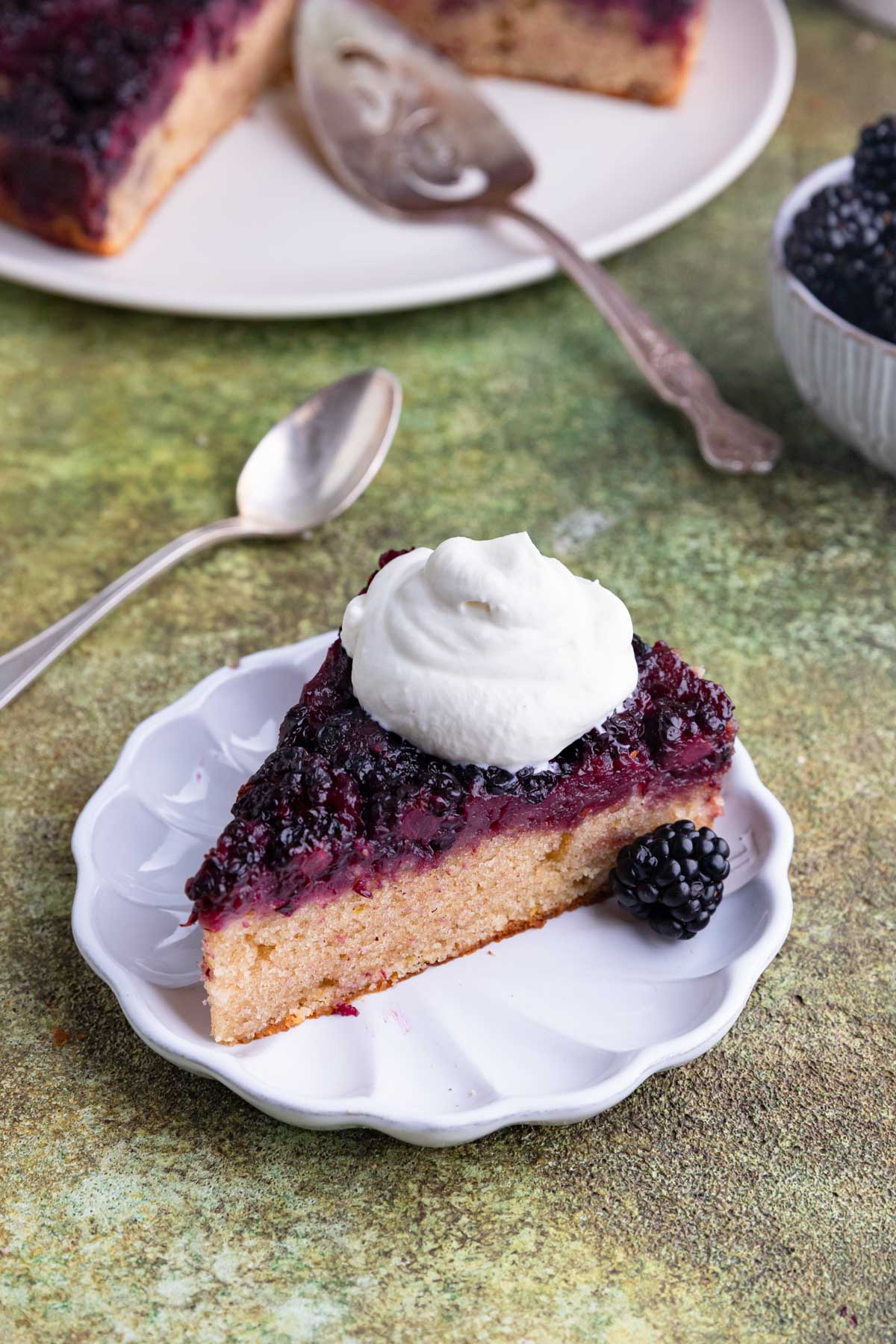 A slice of blackberry upside cake with a dollop of whipped cream on a small white plate. The berries on top are dark purple.