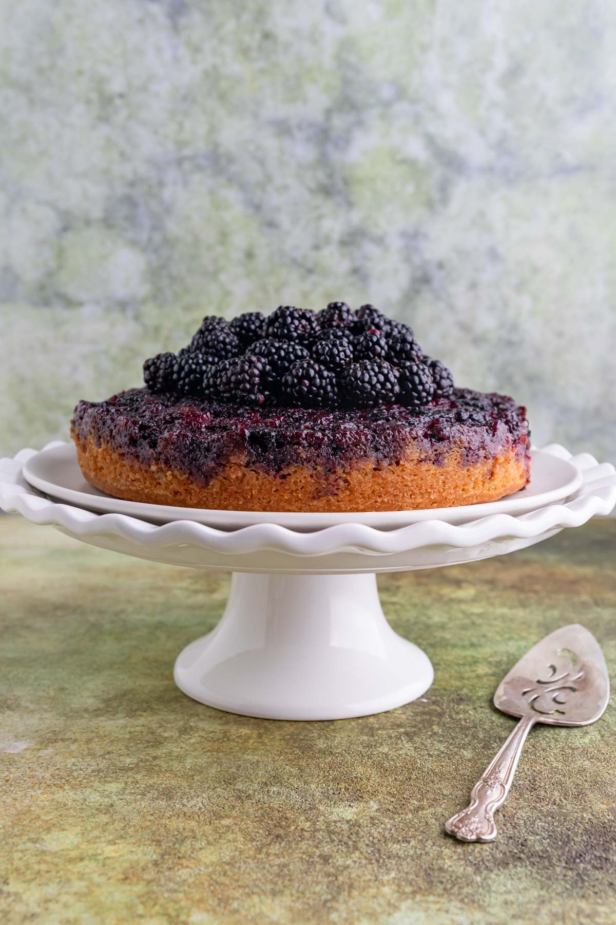 Blackberry upside down cake on a white cake stand with a cake server next to the stand.  The cake has fresh blackberries piled on top