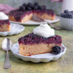 A slice of blackberry upside down cake with whipped on top on a small white plate with the rest of the cake and a bowl of fresh blackberries in the background