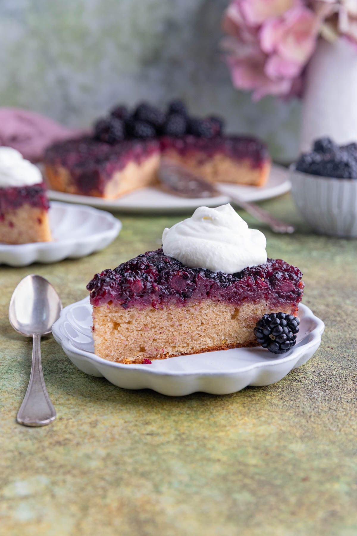 A slice of blackberry upside down cake with whipped on top on a small white plate with the rest of the cake and a bowl of fresh blackberries in the background