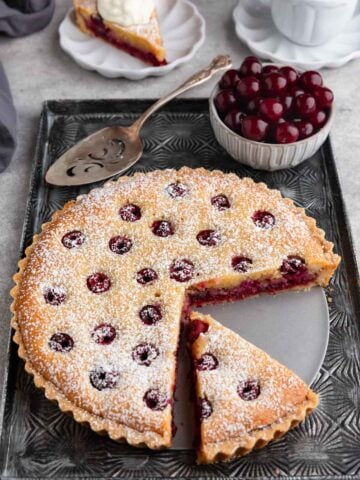 A cherry frangipane tart on a dark gray tray with a bowl of pie cherries next to the tart. Two slices are cut out of the tart. One is on a small plate in the background and the other is on the tart bottom tray