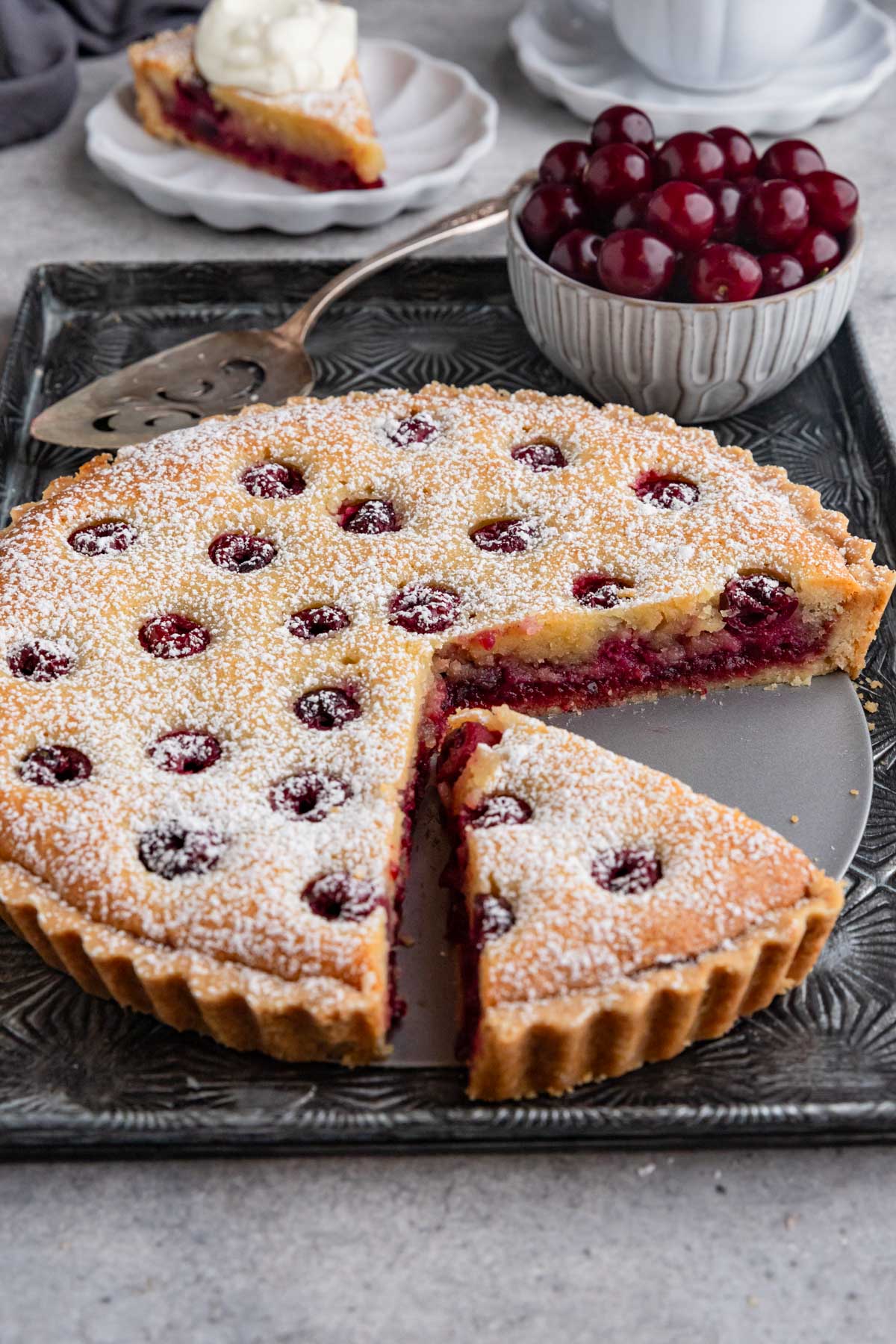 A cherry frangipane tart on a dark gray tray with a bowl of pie cherries next to the tart.  Two slices are cut out of the tart.  One is on a small plate in the background and the other is on the tart bottom tray