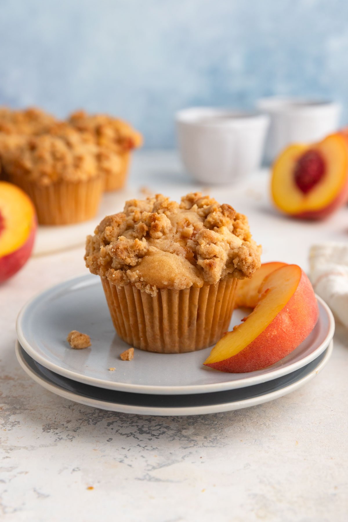 A single peach muffin on a small blue plate with fresh peach slices next to the muffin.  There are more muffins in the background.