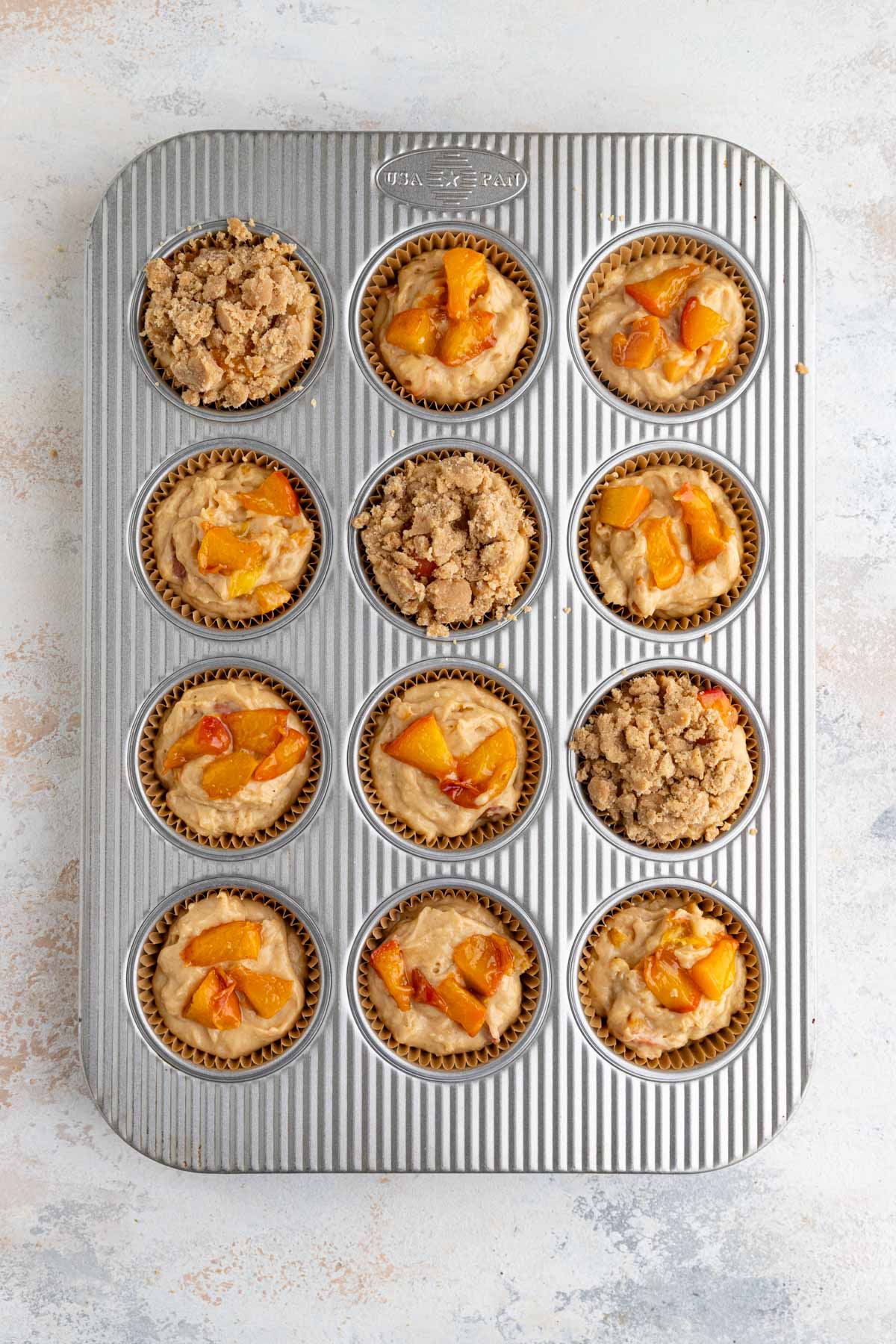 Unbaked peach muffins in a 12 cup muffin tip. Some of the muffins are topped with chunks of peach and others are topped with brown sugar crumble