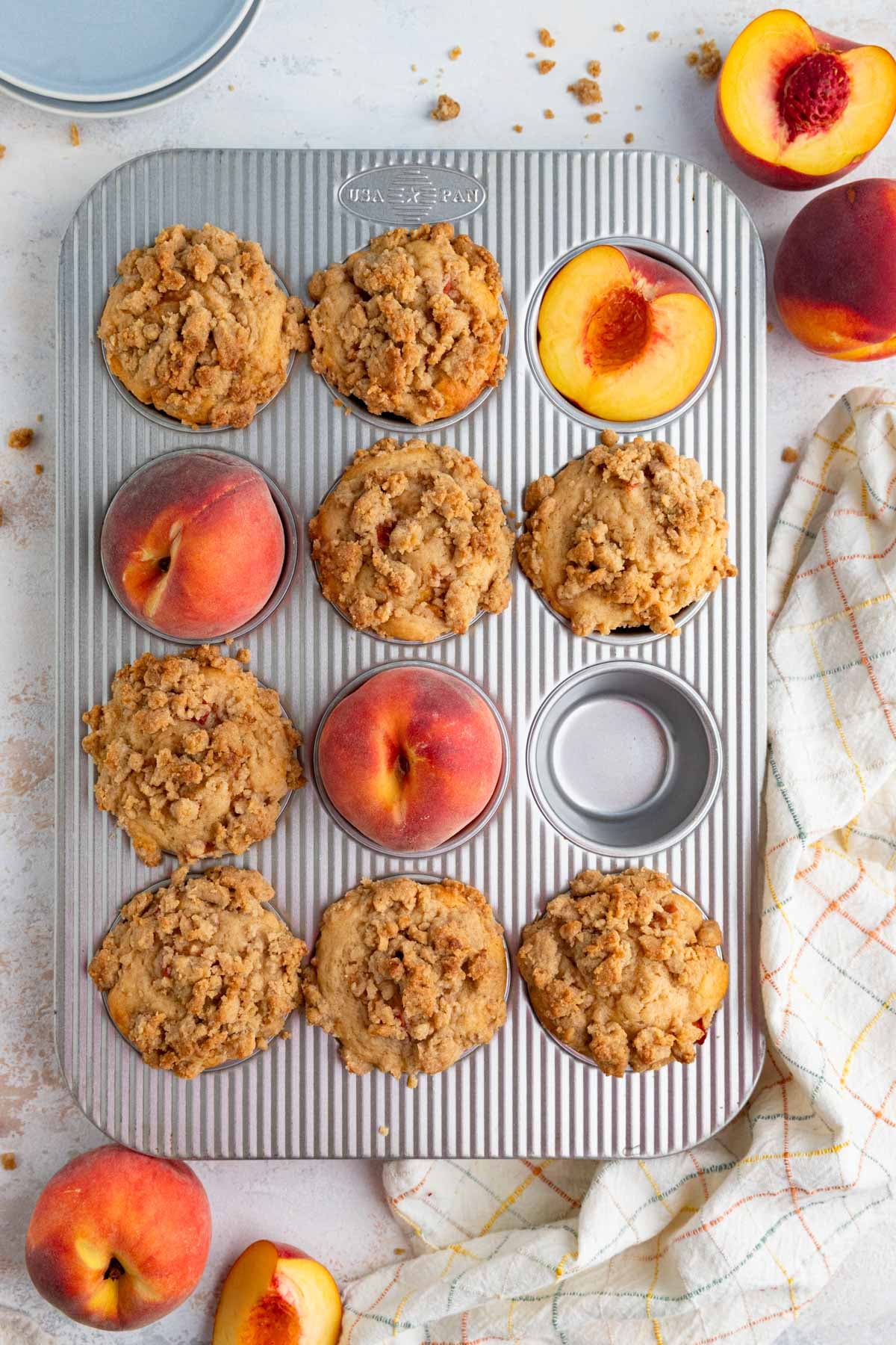 Baked peach streusel muffins in a 12 cup muffin pan. Three of the cups are filled with fresh peach, one is empty and the rest are filled with baked muffins