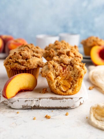 A wooden board with 5 peach streusel muffins and one peach slice. One muffin has bite taken out of it and you see the peaches inside the muffin