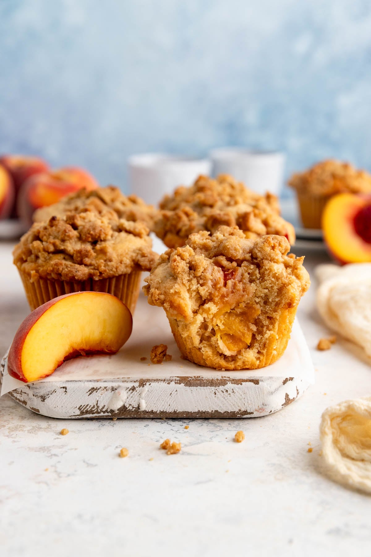 A wooden board with 5 peach streusel muffins and one peach slice. One muffin has bite taken out of it and you see the peaches inside the muffin