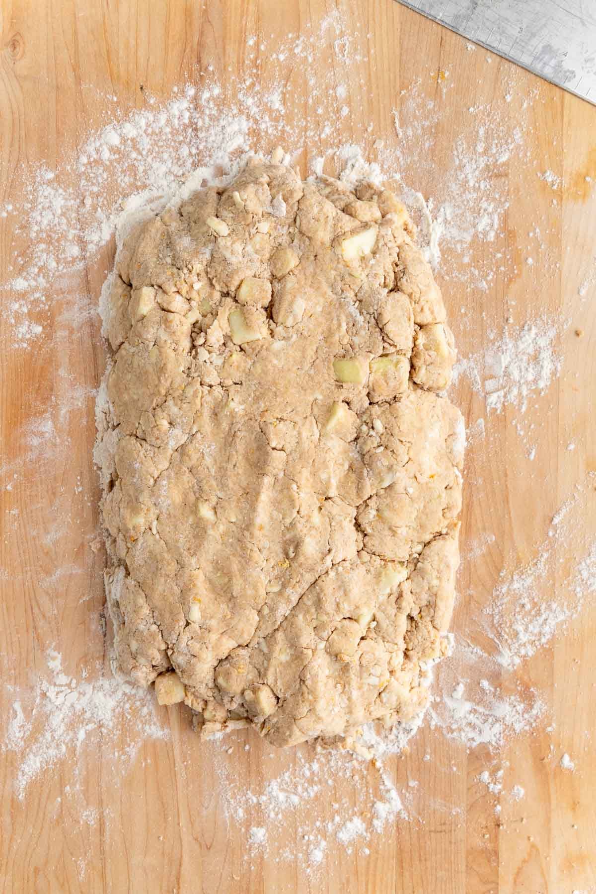The apples scone dough shaped into a rectangle on a well-floured wooden board. You can see chunks of apple throughout the dough.
