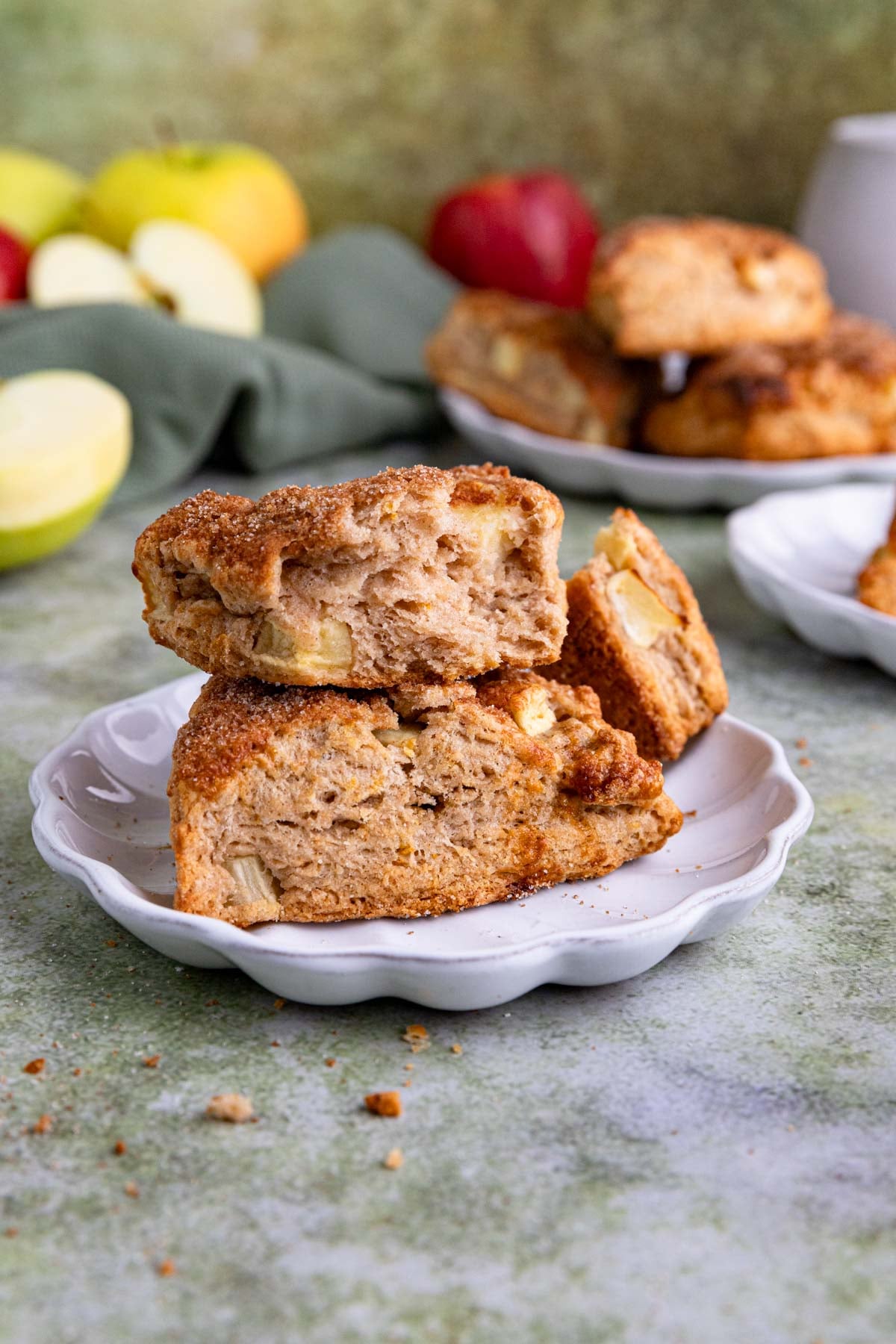 Two apple scones on a white small plate. One of the scones is broken in half. Apple chunk are visible in both scones. There are more scones in the background.