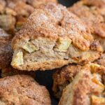 An apple scone piled on top of other apple scones. The scone is triangular, golden brown, and the top is coated with a sugar cinnamon mixture. Chunks of apple are visible.