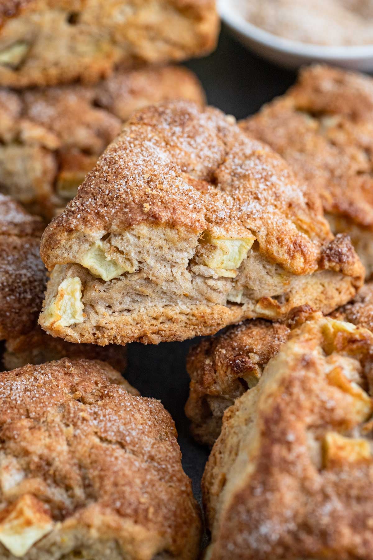 An apple scone piled on top of other apple scones. The scone is triangular, golden brown, and the top is coated with a sugar cinnamon mixture. Chunks of apple are visible.