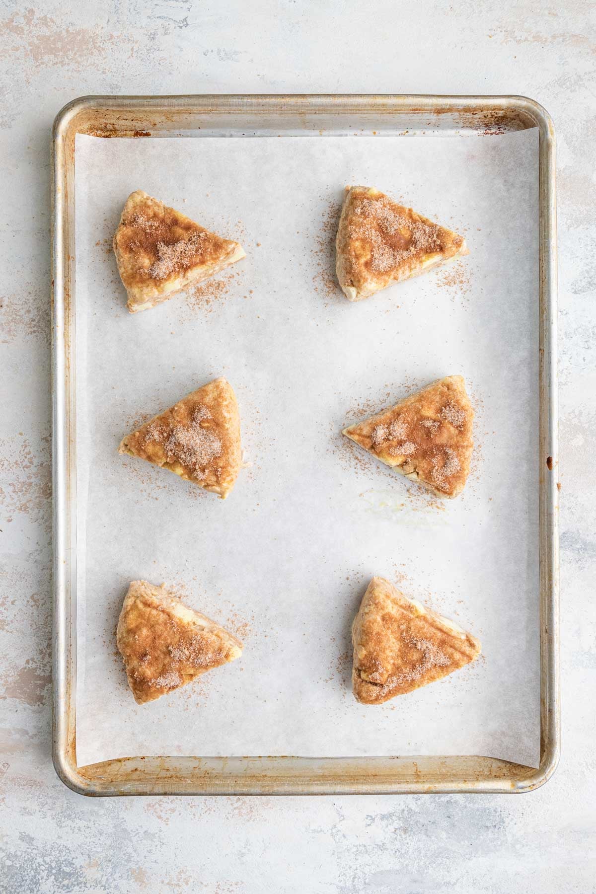 Six unbaked triangular shaped scones on a parchment lined baking sheet. Each scone is heavily coated with a sugar cinnamon mixture