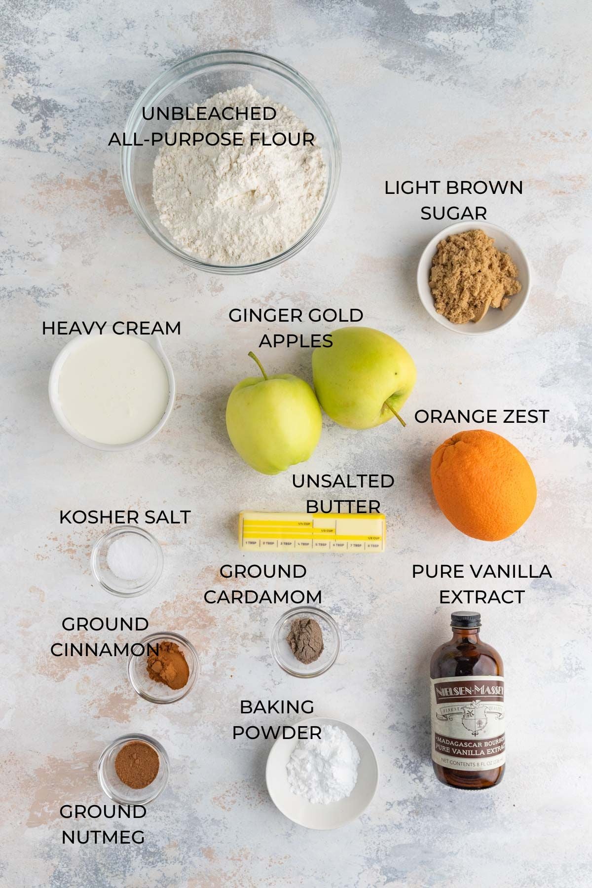 Apple scone ingredients - all purpose flour, light brown sugar, a whole orange, pure vanilla extract, baking powder, ground cardamom, ground cinnamon, unsalted butter, kosher salt, Ginger Gold apples, and heavy cream on a board