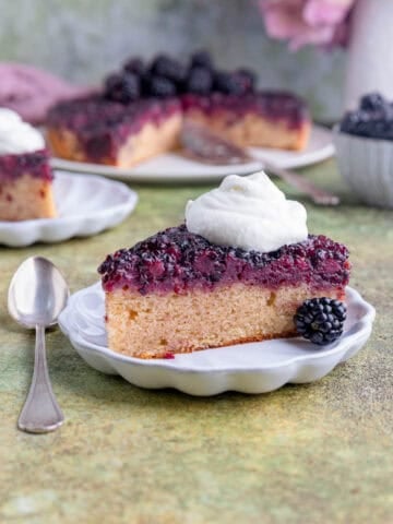 A slice of blackberry upside down cake with whipped on top on a small white plate with the rest of the cake and a bowl of fresh blackberries in the background