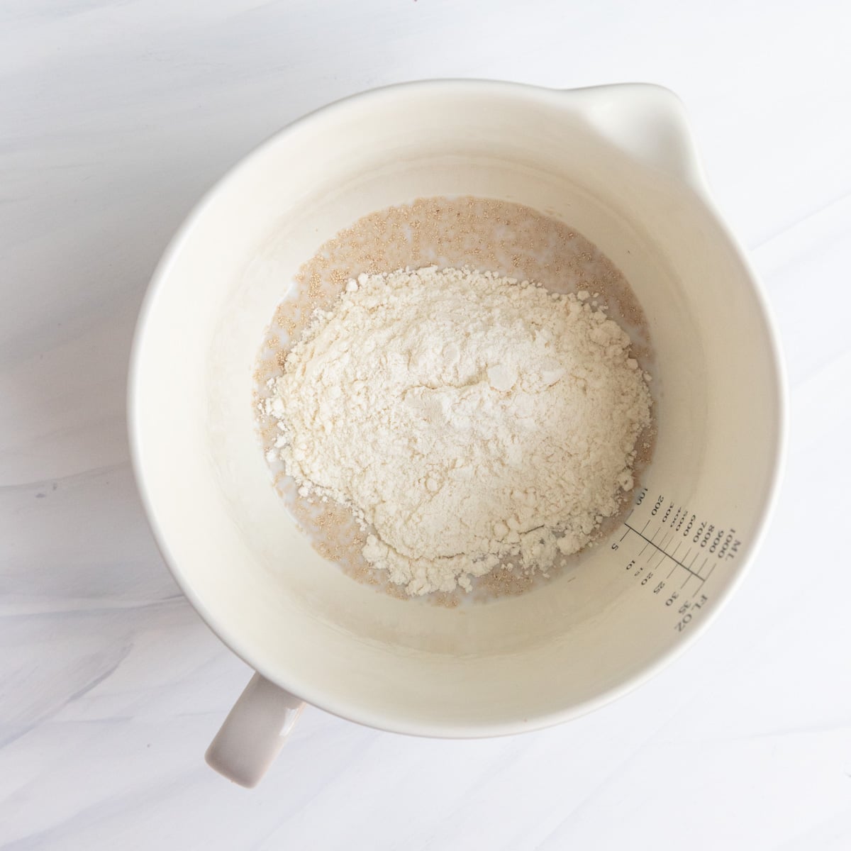All-purpose flour sprinkled on top of a yeast, sugar, and milk mixture in a ceramic bowl